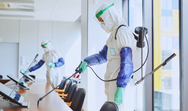 Professionals in full protective gear performing disinfection services in a modern office, using spray equipment to sanitize surfaces.