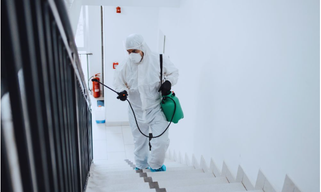 A professional in full protective gear using a sprayer to disinfect a stairwell in a residential building.