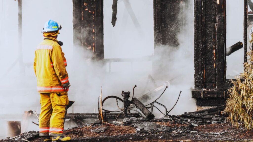 A fire damage restoration professional in yellow protective gear and a blue helmet stands in front of a burned, smoky building, assessing the damage, with charred debris and structural remains visible nearby.