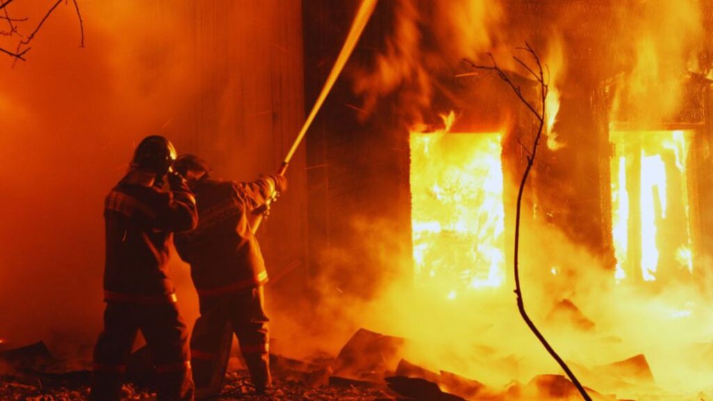 Two firefighters in protective uniforms spray water on a burning building, surrounded by intense flames and thick smoke, illustrating emergency fire damage restoration near me services in action.